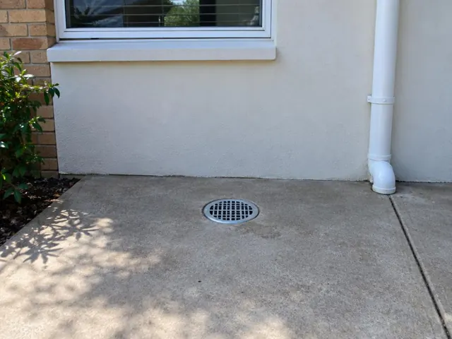 Outdoor gully grate set in concrete paving beside a house wall and downpipe in Sutherland