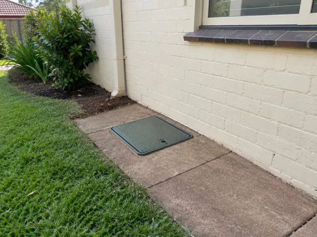 Flush green inspection cover set into a concrete path beside a cream brick house and lawn in Gymea Bay